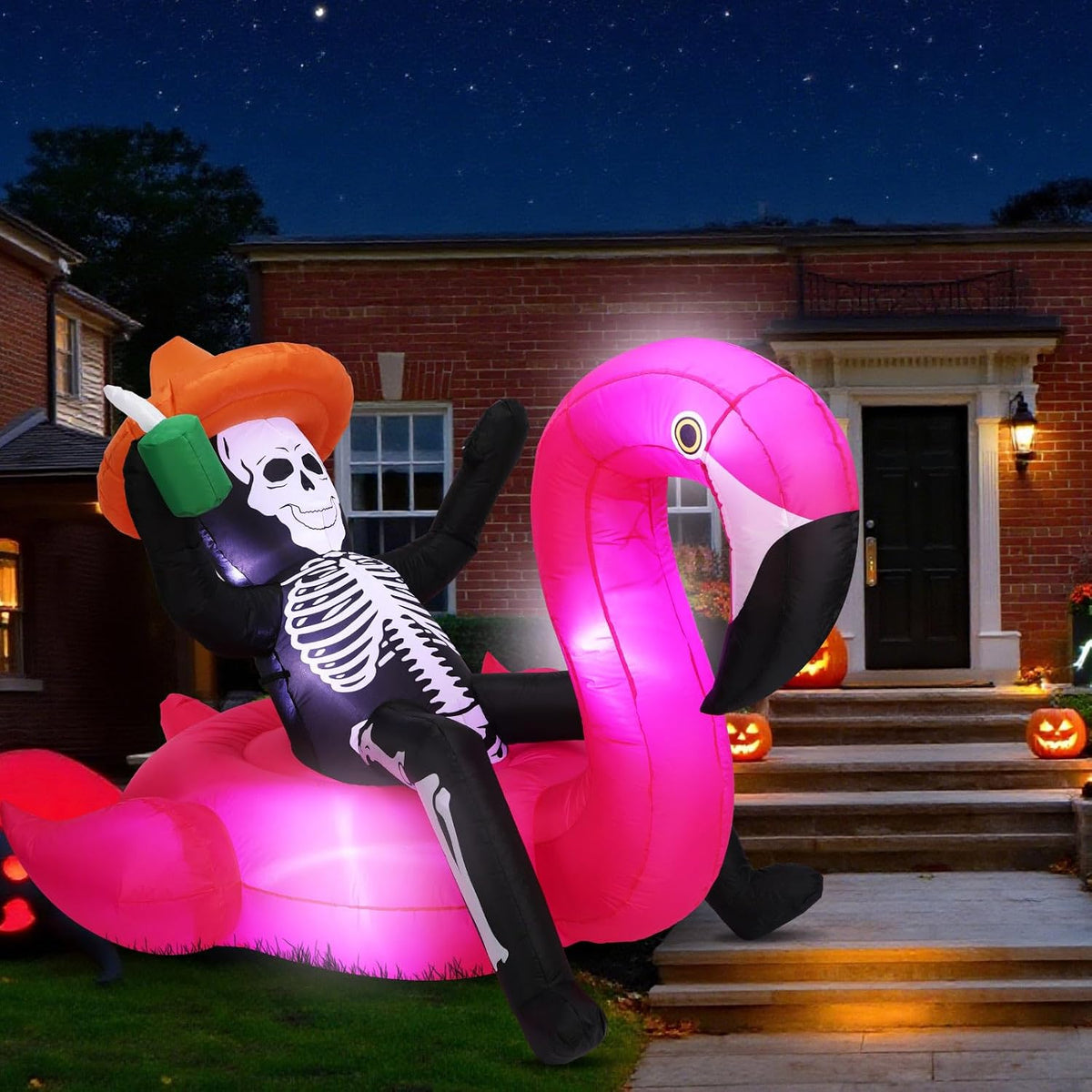 Inflatable skeleton in an orange hat lounging on a pink flamingo in front of a house at night