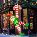 Elf inflatable in green with striped legs hanging from a giant candy cane on the front of a building