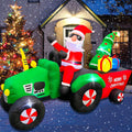 Santa in sunglasses driving a green tractor pulling a red Merry Christmas wagon with a tree and gifts