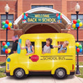 Yellow inflatable school bus with kids leaning out of the windows in front of a school with a Welcome Back to School sign